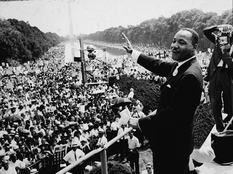 A black man dressed in a dark suit waves before speaking to thousands of people gathered around the Lincoln Memorial.