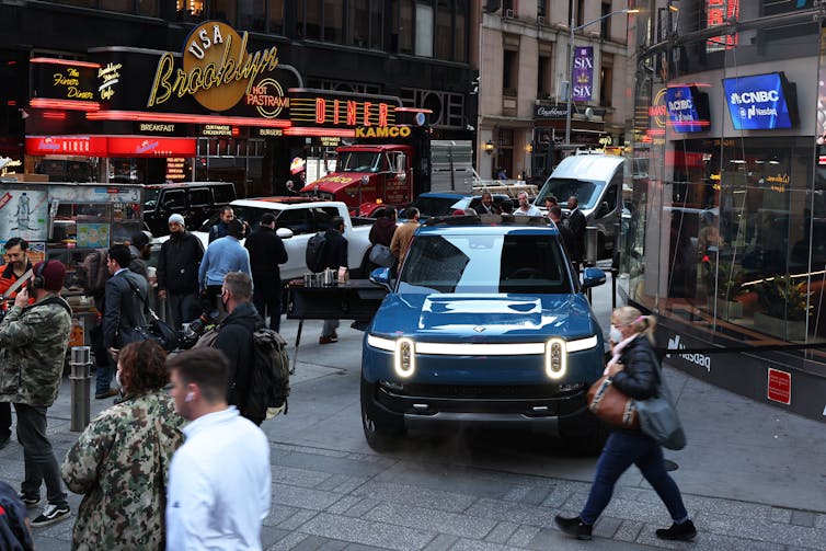 A Rivian EV pickup sits in front of a TV studio in New York with people walking around it.
