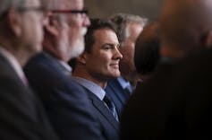 A dark-haired man smiles among other men with grey hair.