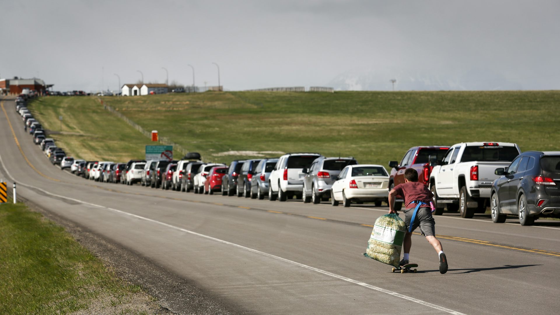 A teenaged boy on a skateboard holds bags of popcorn as he moves along a long row of cars on a highway.
