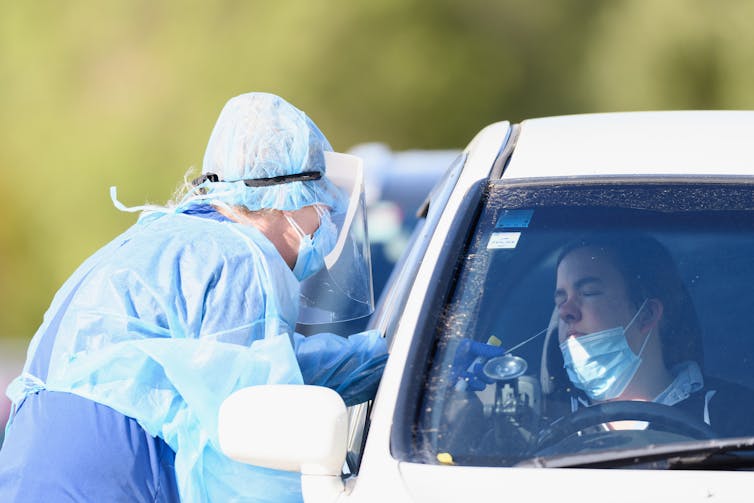 Nurse in protective gear and mask leans into car to swab driver.