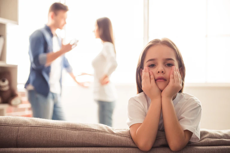 A sad little girl looks into the camera while her parents argue behind her