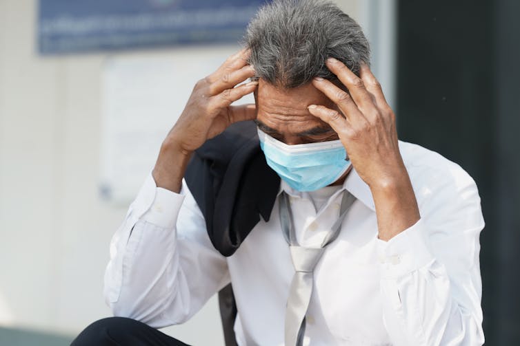 A business man wearing a face mask looks sad and rests his head on his hands