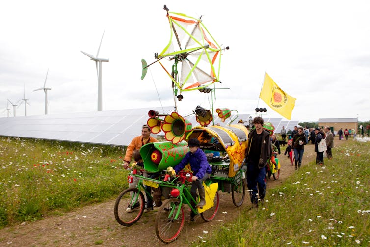 A procession of people with flags march past solar panels and wind turbines.