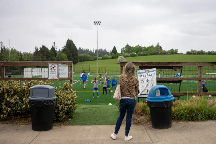 A woman in a leoplard print jacket seen by herself near a covid sign watching a soccer game.