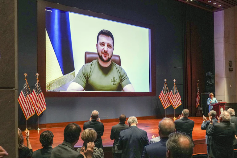 A man on a screen displayed above a stage set with American flags. Other people facing the screen stand clapping.