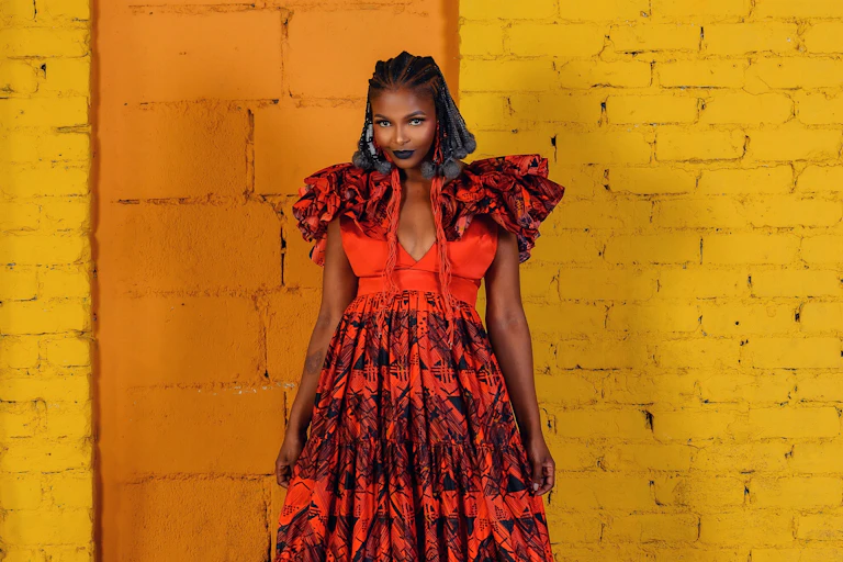 A woman stands against a brick wall, painted orange and yellow. She wears a red and black dress and has blue and black hair in thick braids, a slight smile on her face.