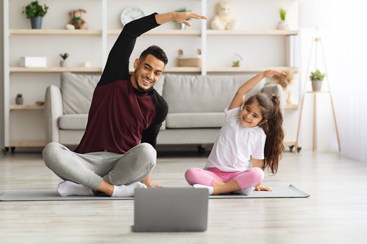 A father and daughter seen in front of a laptop doing exercise on the floor.