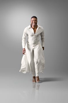 A man strides towards the camera with a big smile on his face. He wears a white one-piece outfit, the top like a dress-up shirt and the bottom a flowing skirt.