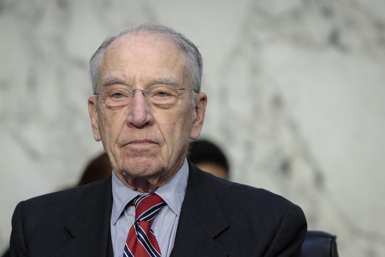 An elderly white man wearing a dark business suit is seen with a marble wall in the background.