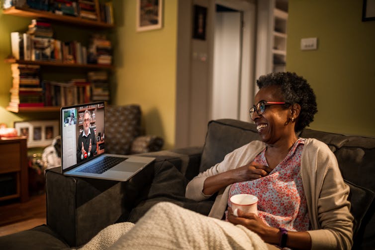 An older Black woman sits on a sofa in her living room, looking at a laptop next to her, where her therapist is on the screen.
