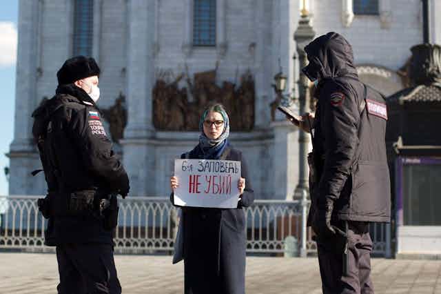 A woman in a headscarf and black coat stands holding a sign in front of a church.