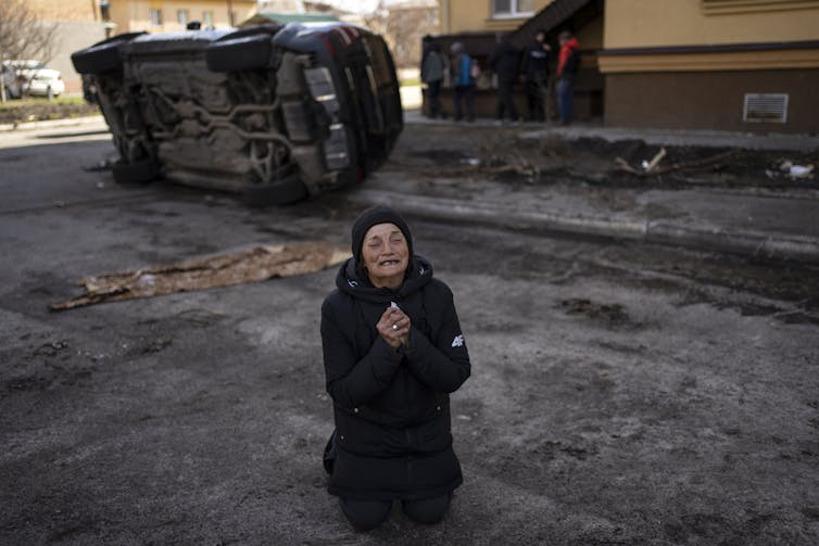 A woman dressed in black kneels on the ground in front of an overturned truck and weeps.