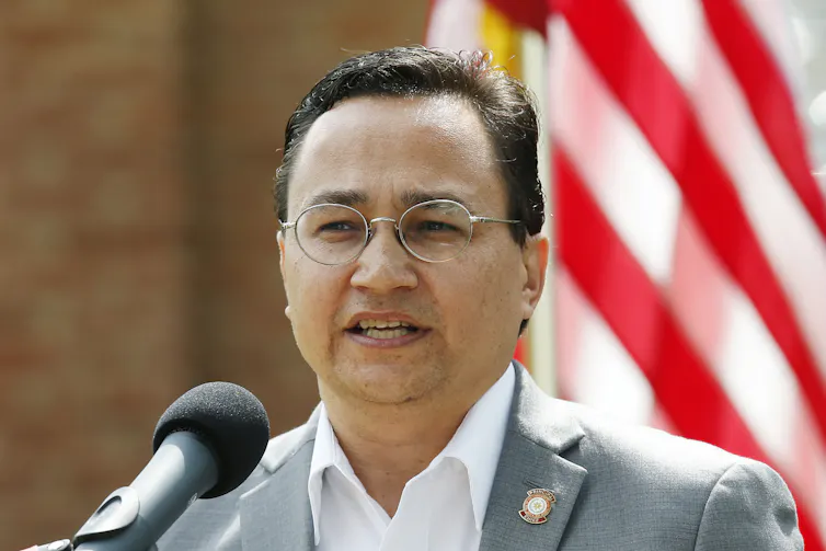 A man in a coat and collared shirt stands in front of a microphone.