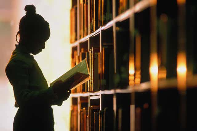 A young girl stands in front of a bookshelf with a book in her hands.