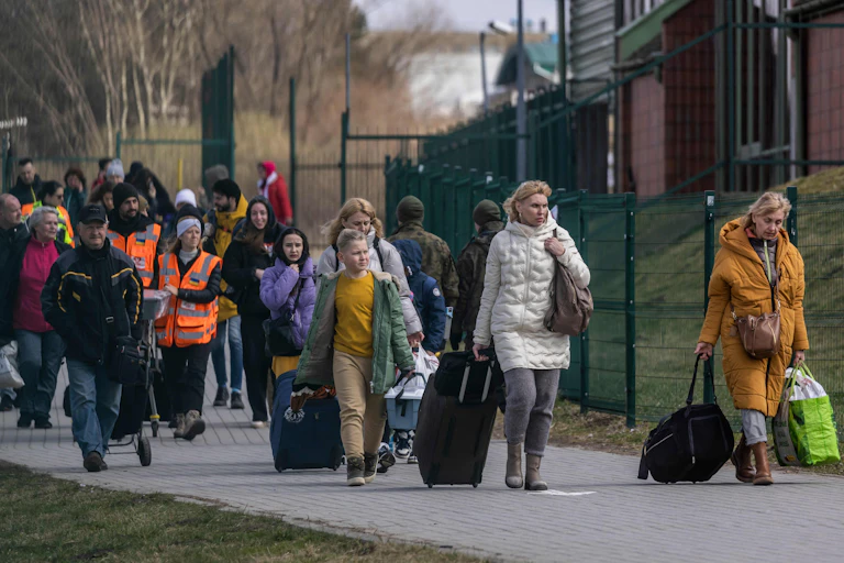 A group of people, including two women at the front, wheeling suitcases are seen walking on a sidewalk.