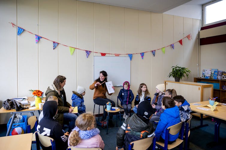 Children surround an adult in a classroom, all seated, with a rainbow flag hanging on a wall.