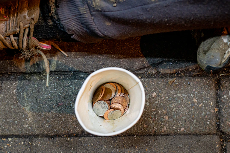 Photo from above of some change in a paper cup in front of a person sitting on the sidewalk