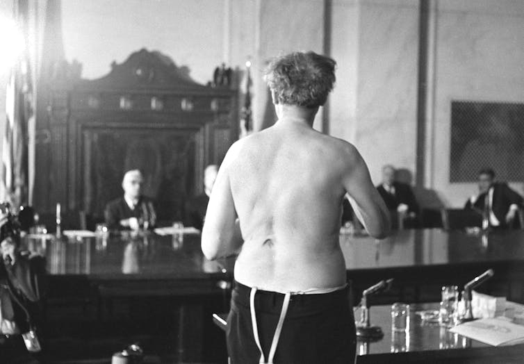 A man, stripped to the waist, showing scar marks on his back to a committee seated in front of him