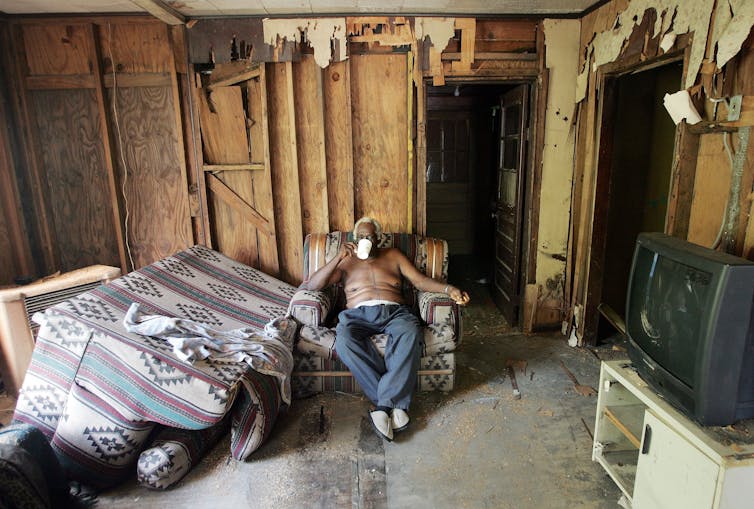 An older Black man sits in a seat drinking from a mug, in a dilapidated looking room with exposed wooden beams and walls.