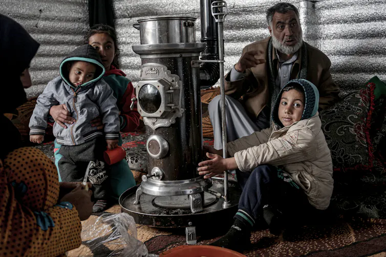 A woman and man sit in front of two young children in a tent, next to a large heater.
