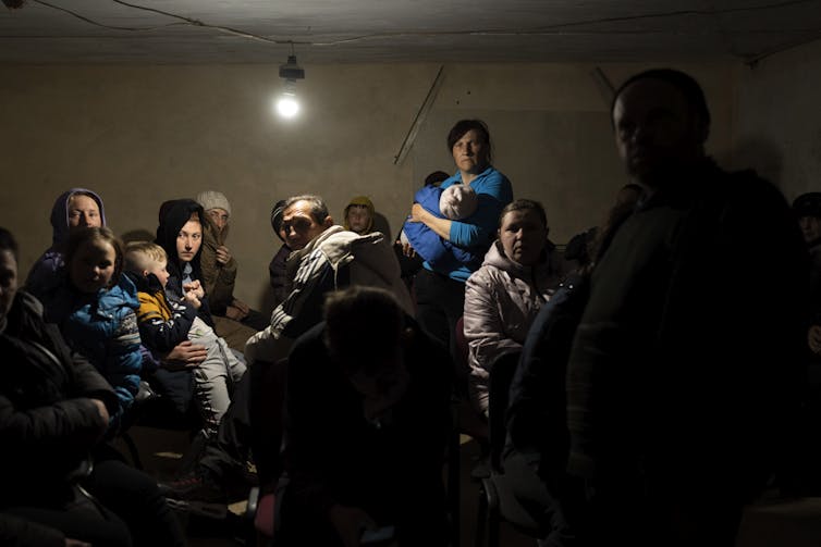 Men, women and children huddle in a basement under a single light bulb.