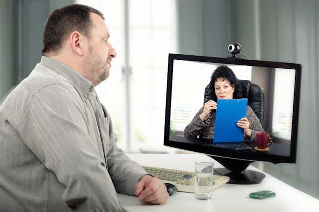 Large-bodied white man looks at a computer screen with a camera on top of it pointed at him. On the screen is a white woman holding a blue clipboard.