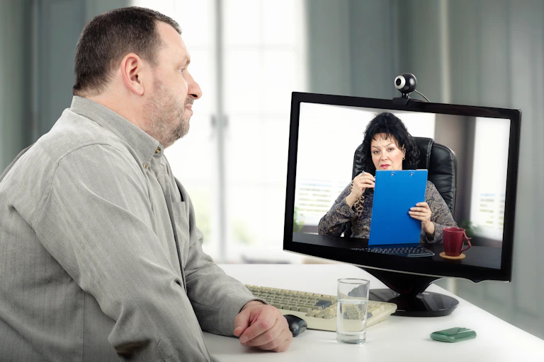 Large-bodied white man looks at a computer screen with a camera on top of it pointed at him. On the screen is a white woman holding a blue clipboard.