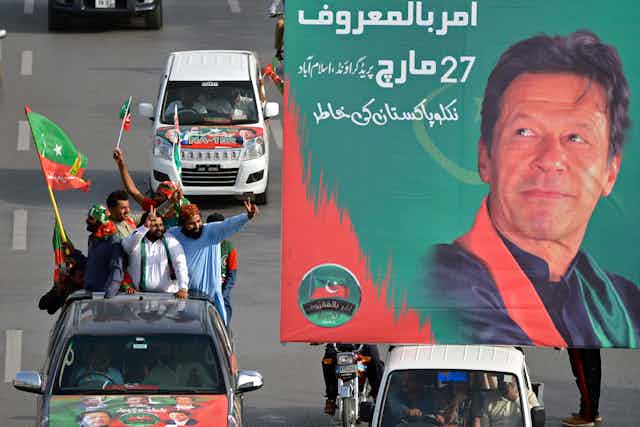 Men in a truck waving flags gesture towsrds a poster showing Imran Khan.