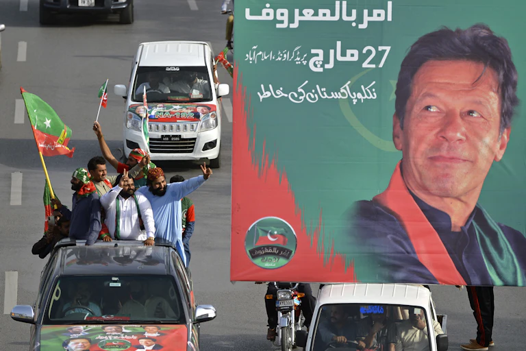 Men in a truck waving flags gesture towsrds a poster showing Imran Khan.