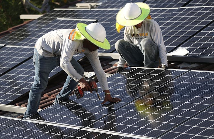 Two workers in wide-rimmed hats install solar panels on a roof