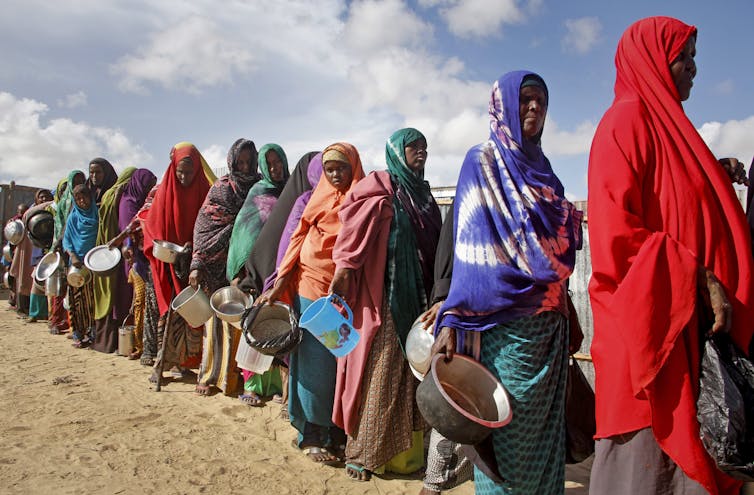 Women stand in a queue with empty food containers.