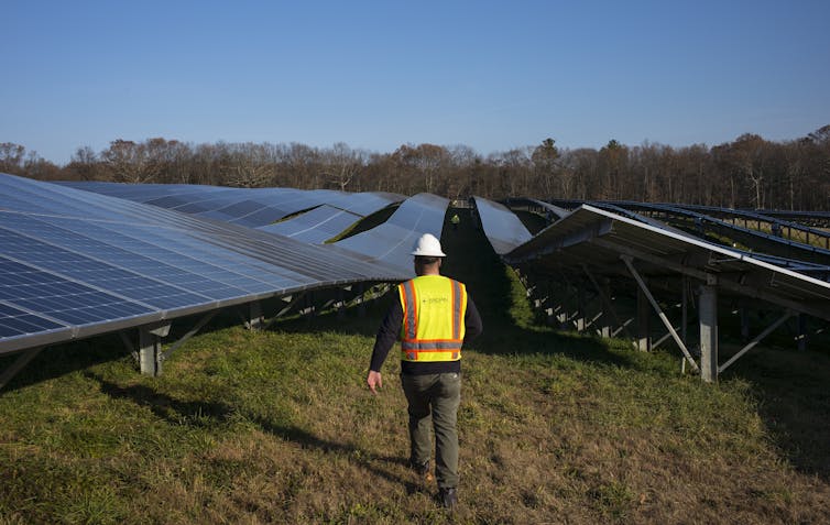 A worker in a hard hat and construction vest walks between rows of solar panels in a field.
