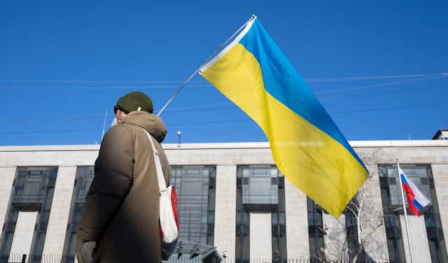 A person in a brown winter coat holds the Ukrainian flag as they walk past a building with a Russian flag flying in front of it.