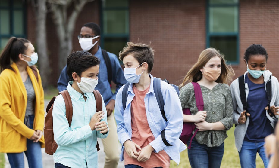 A multi-ethnic group six of middle school students walking outdoors, in front of the school building, carrying backpacks. They are back to school during the COVID-19 pandemic, wearing protective face masks.