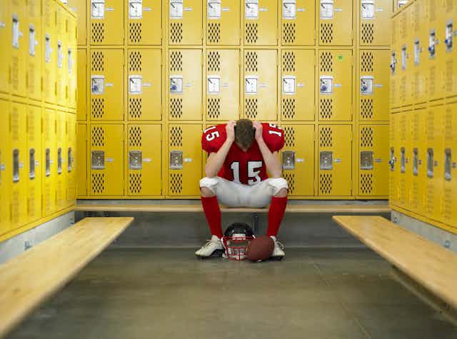 A high school football player, wearing a red shirt with the number 15 in white and white pants, sits alone on a bench in a locker room with a football helmet and football on the floor.
