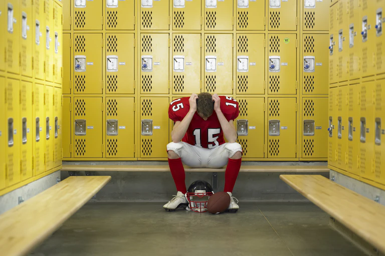 A high school football player, wearing a red shirt with the number 15 in white and white pants, sits alone on a bench in a locker room with a football helmet and football on the floor.