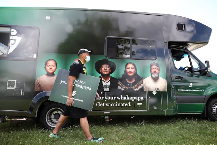 Man with signs walks past a caravan used for vaccinations.