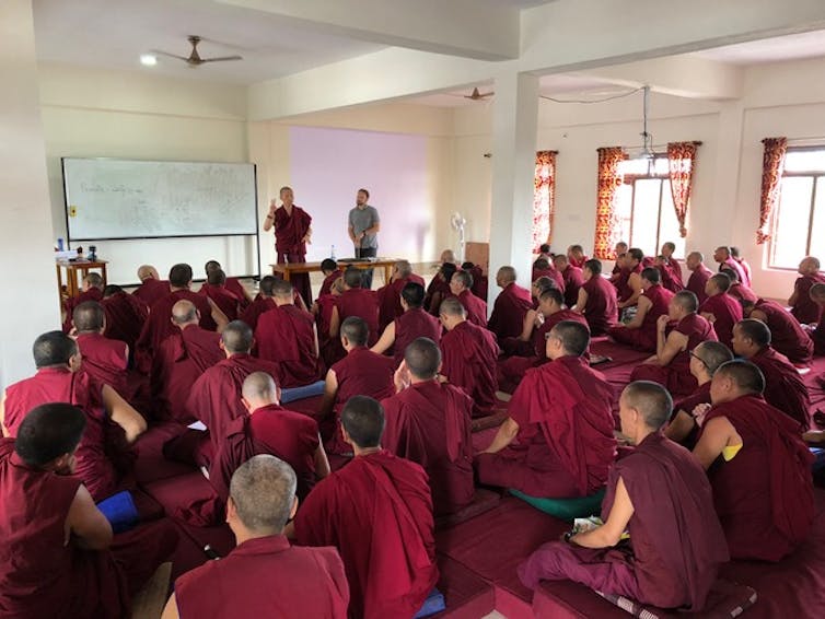 Monks in red robes sit in a classroom as two teachers talk at the front of the room.