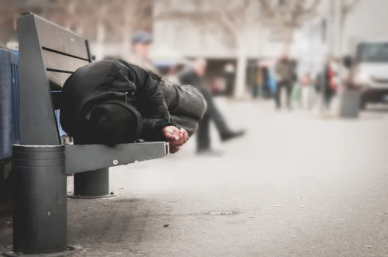 Homeless man sitting on a public bench, hunched over.