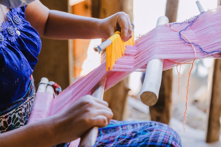A woman weaves pink and yellow yarns into frabric using wooden poles.