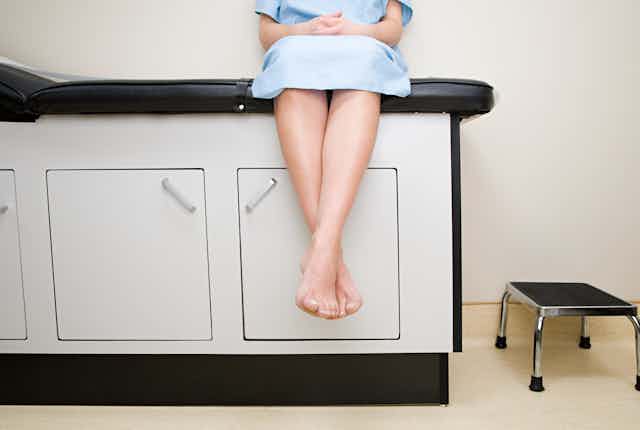 Cropped image showing a person from the chest down sitting on a medical exam table wearing a hospital gown