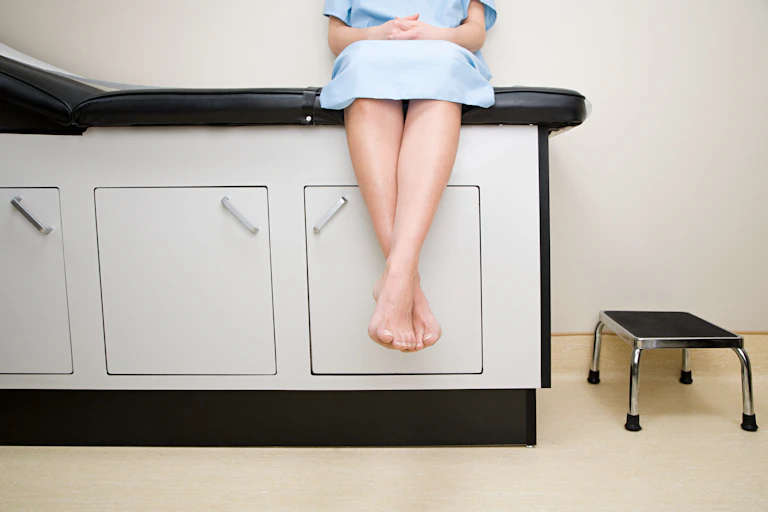 Cropped image showing a person from the chest down sitting on a medical exam table wearing a hospital gown