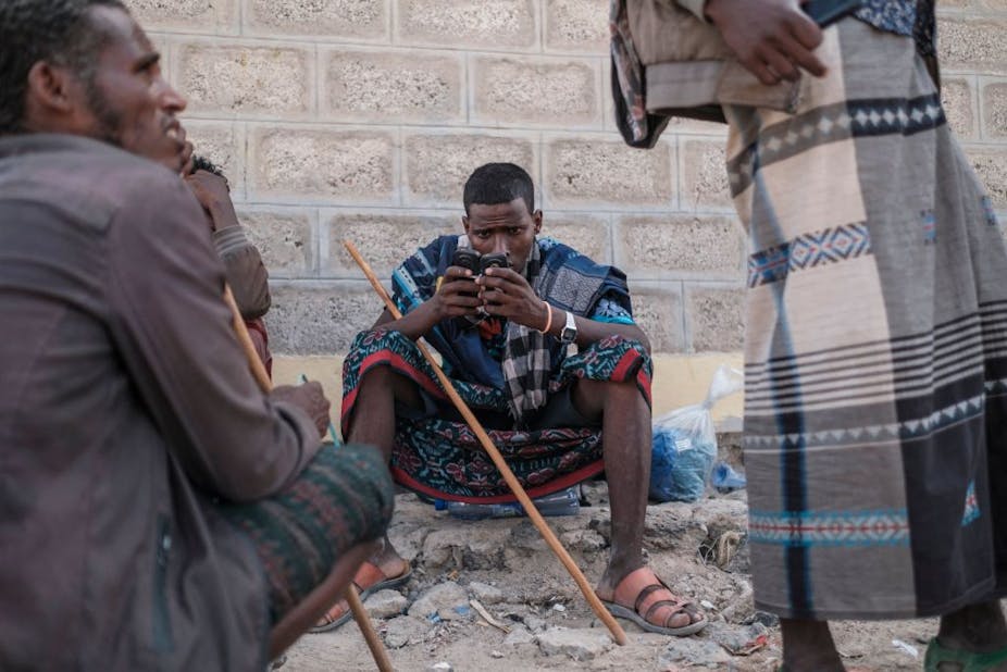 A man sits outside a building, holding a mobile phone. He and another man in the picture carry sticks.