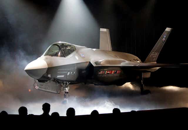 A fighter jet sit on a stage under spotlights and enshrouded in dry ice.