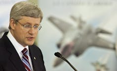A man with grey hair and glasses speaks into a microphone with a photo of a fighter jet in the background.