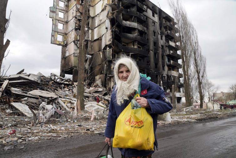 An elderly woman with a white wool scarf on her head and carrying a yellow plastic bag walks past a destroyed apartment building.