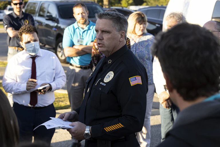 A man in a police uniform speaks to people holding cameras and notebooks