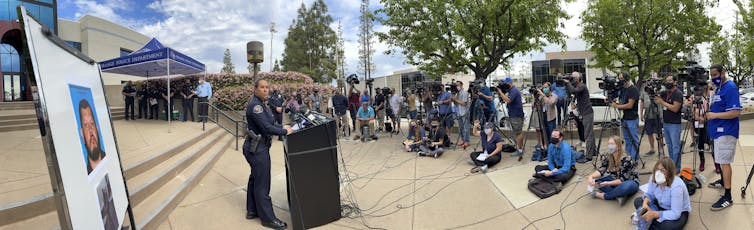 A police officer stands at a lectern facing people holding microphones and cameras