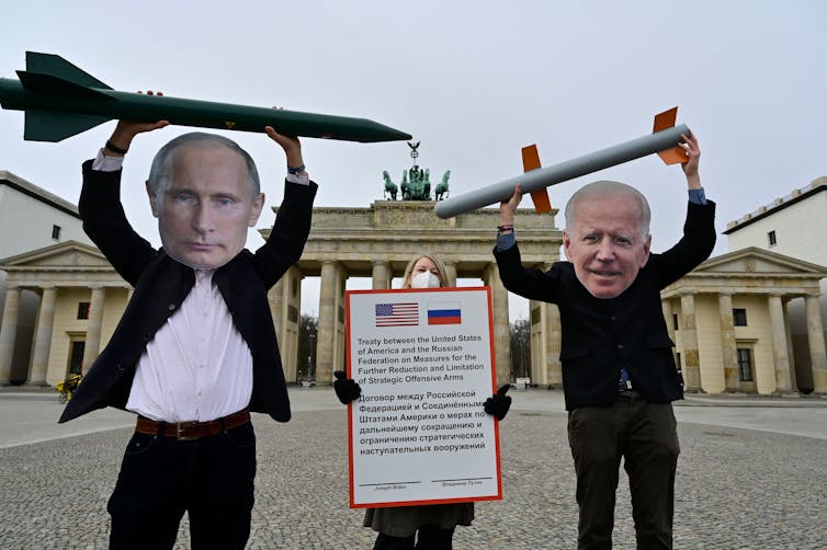 Two people wearing suits and large cut out faces of Putin and Biden hold fake ballistic missiles high above their heads.